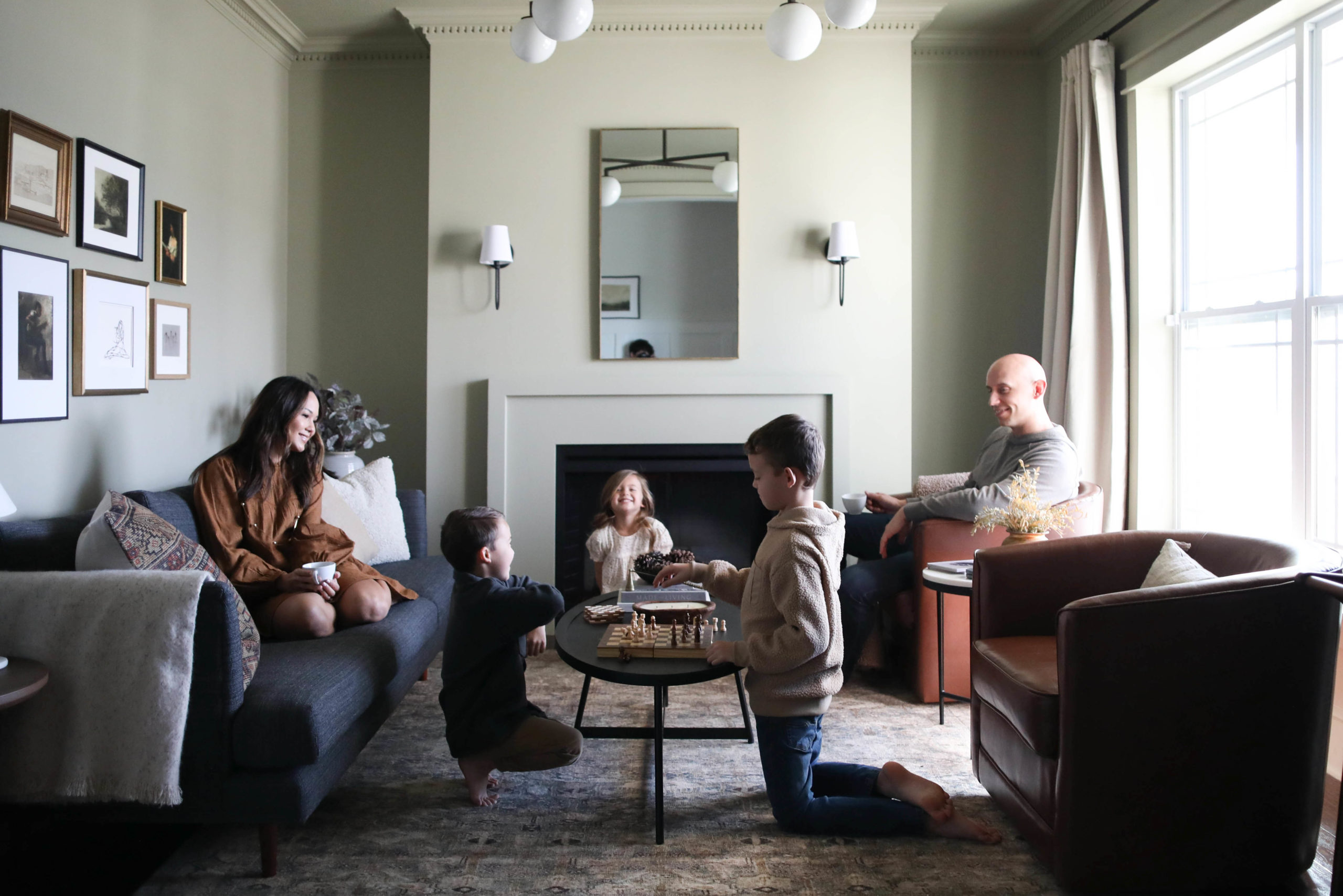 mrsjessicadarling, her husband and three kids are hanging out in their muted green space. The boys are playing chess, the little girl is smiling playfully on the ground, while Jessica and Jeff are smiling and enjoying coffee on a sofa and accent chair.