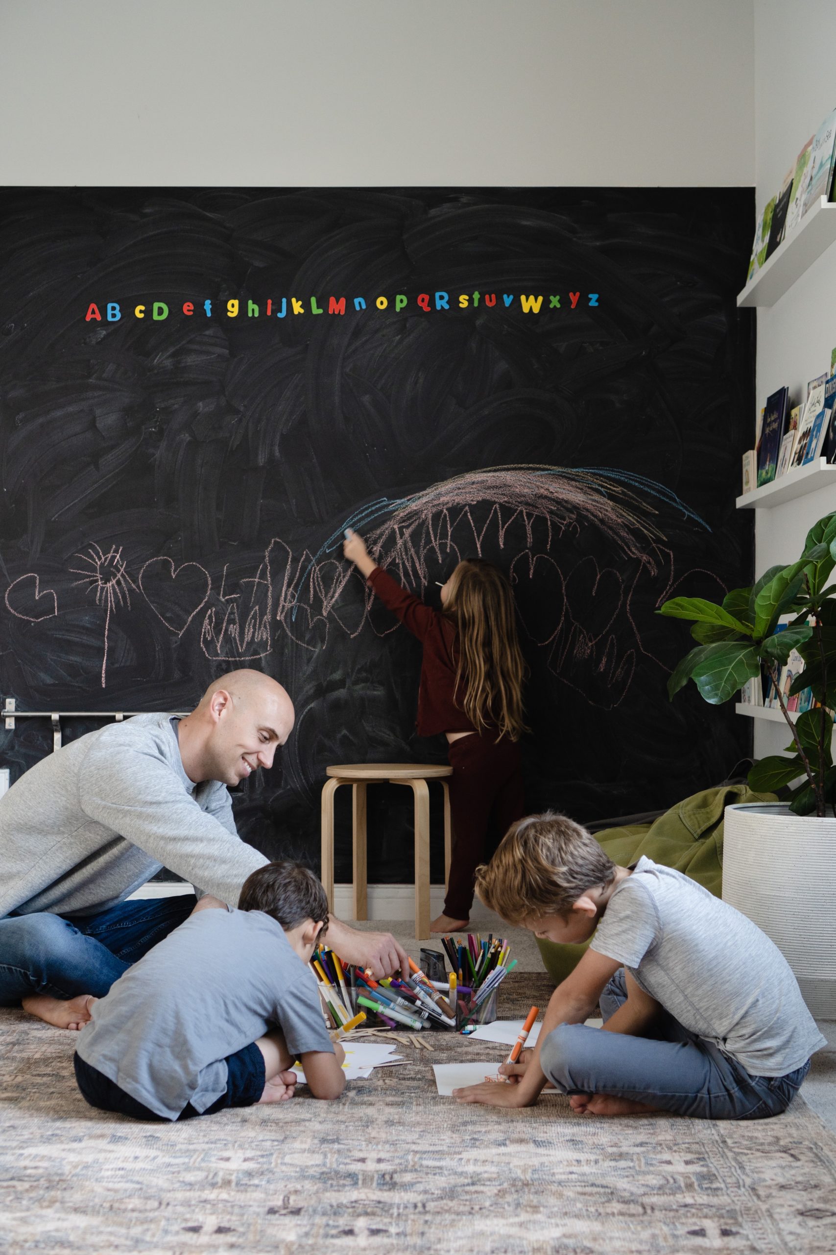 Playroom ideas, with a large magnetic chalkboard wall, featuring The Akemon family coloring and hanging out together on a cozy rug. Shows the space where the new bonus room with feature a window seat, DIY home built-in library,